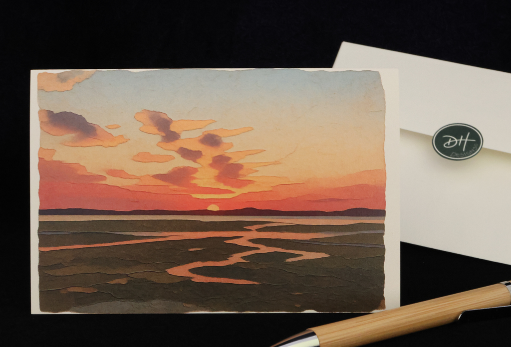 A notecard featuring a sunset landscape with a winding estuary near Morro bay, CA, shown on a black background with a white envelope and a wooden pen nearby.
