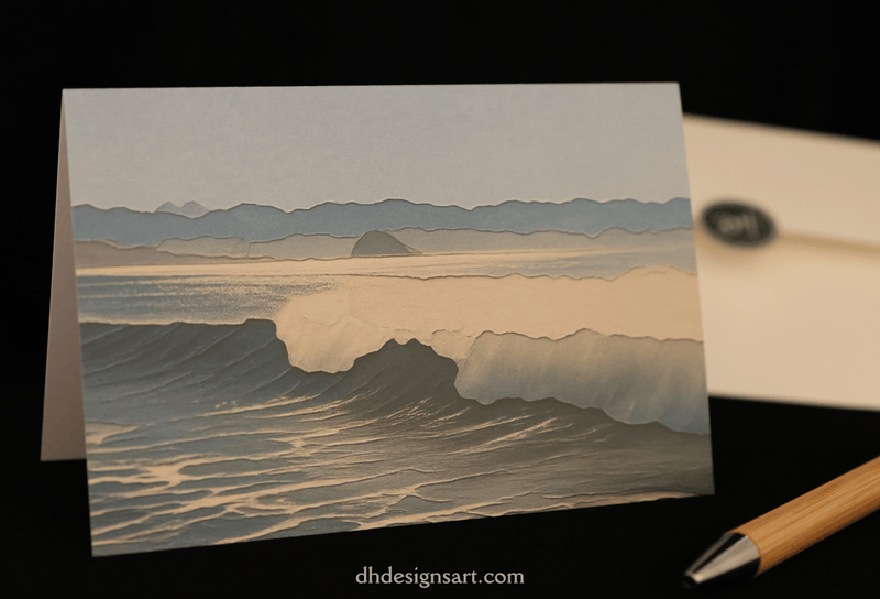 A folded notecard featuring a stylized landscape illustration of Cayucos, CA, with layered mountains, mist, and waves in muted blue, and white tones and Morro Rock in the distance, displayed on a dark surface with an envelope and pen nearby.