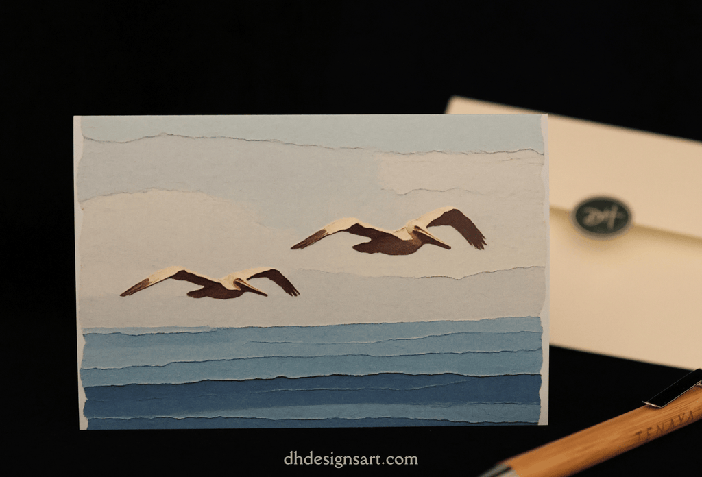A noteard featuring a paper art styled image depicting two seagulls flying over blue ocean waves under a pale sky, displayed next to a cream envelope on a black background.