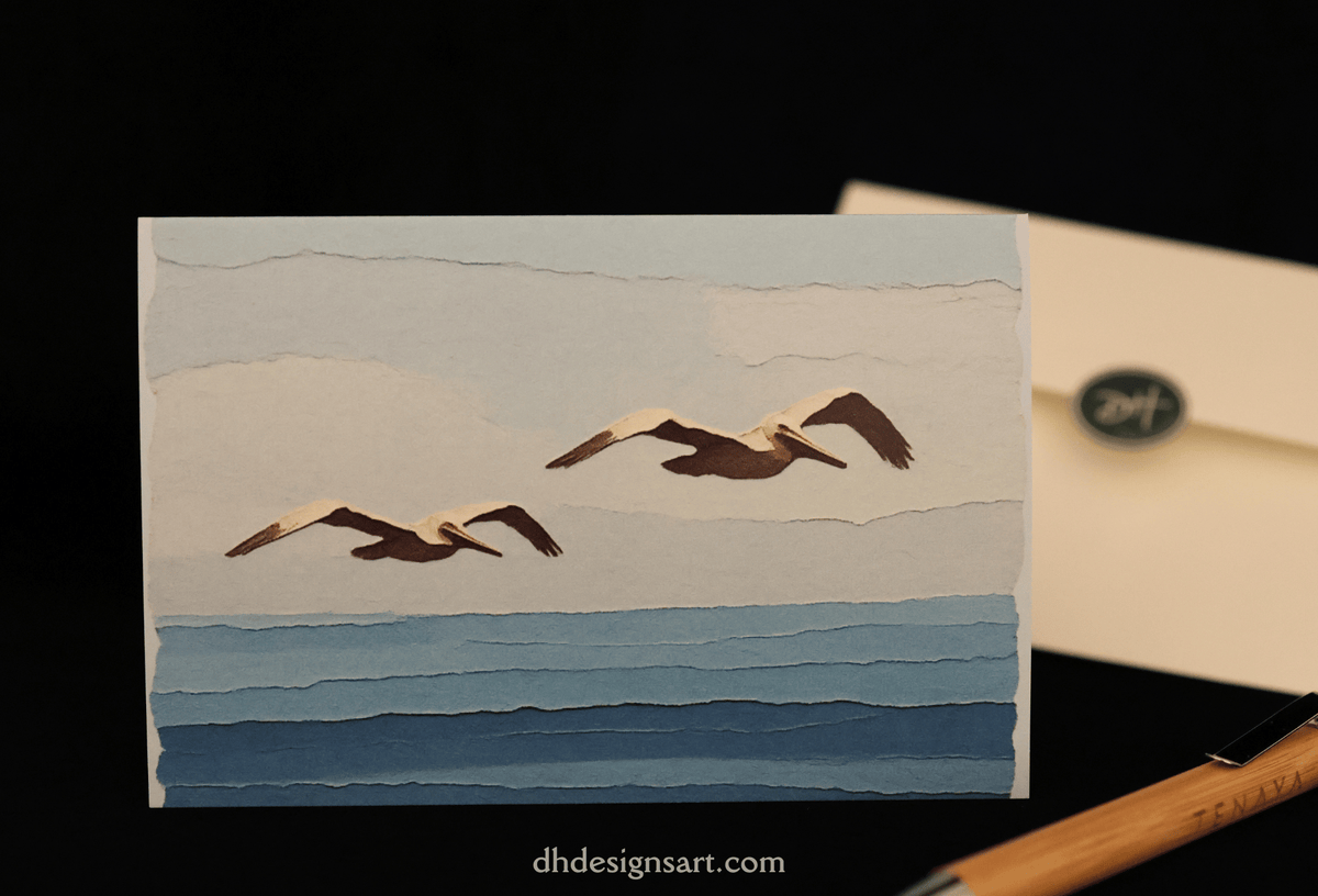 A noteard featuring a paper art styled image depicting two seagulls flying over blue ocean waves under a pale sky, displayed next to a cream envelope on a black background.