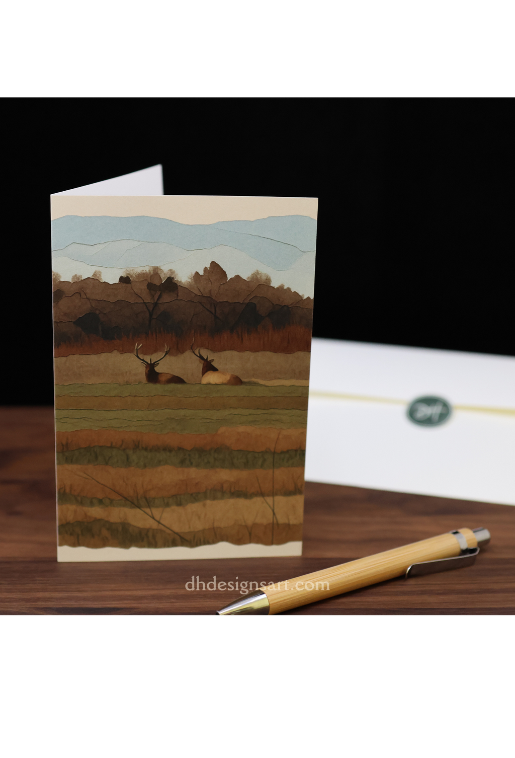 Blank notecard with two Tulle elk on the cover and a pen and envelope on a wooden desk.