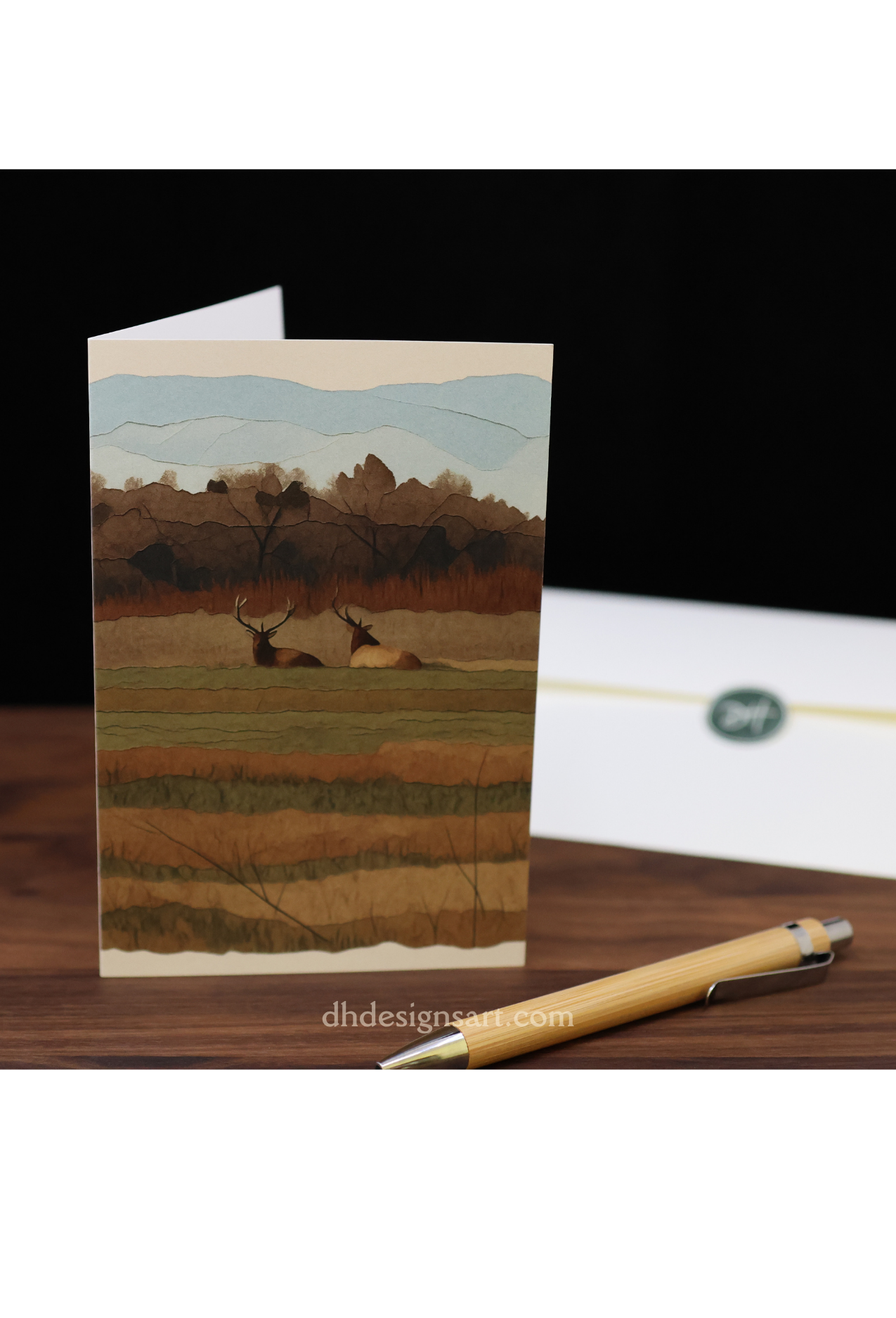 Blank notecard with two Tulle elk on the cover and a pen and envelope on a wooden desk.