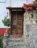 A rustic double wooden door in Tapalpa, Mexico, set in a white and red stucco wall, with a stone wall on the right adorned with bright pink flowers and a potted tree on the left.
