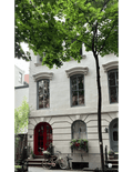 A white stone townhouse facade in Brooklyn, New York, with gray-framed windows, a red arched front door, a blue-gray side door, flower boxes, a bicycle at the entrance, and leafy green trees in the foreground.