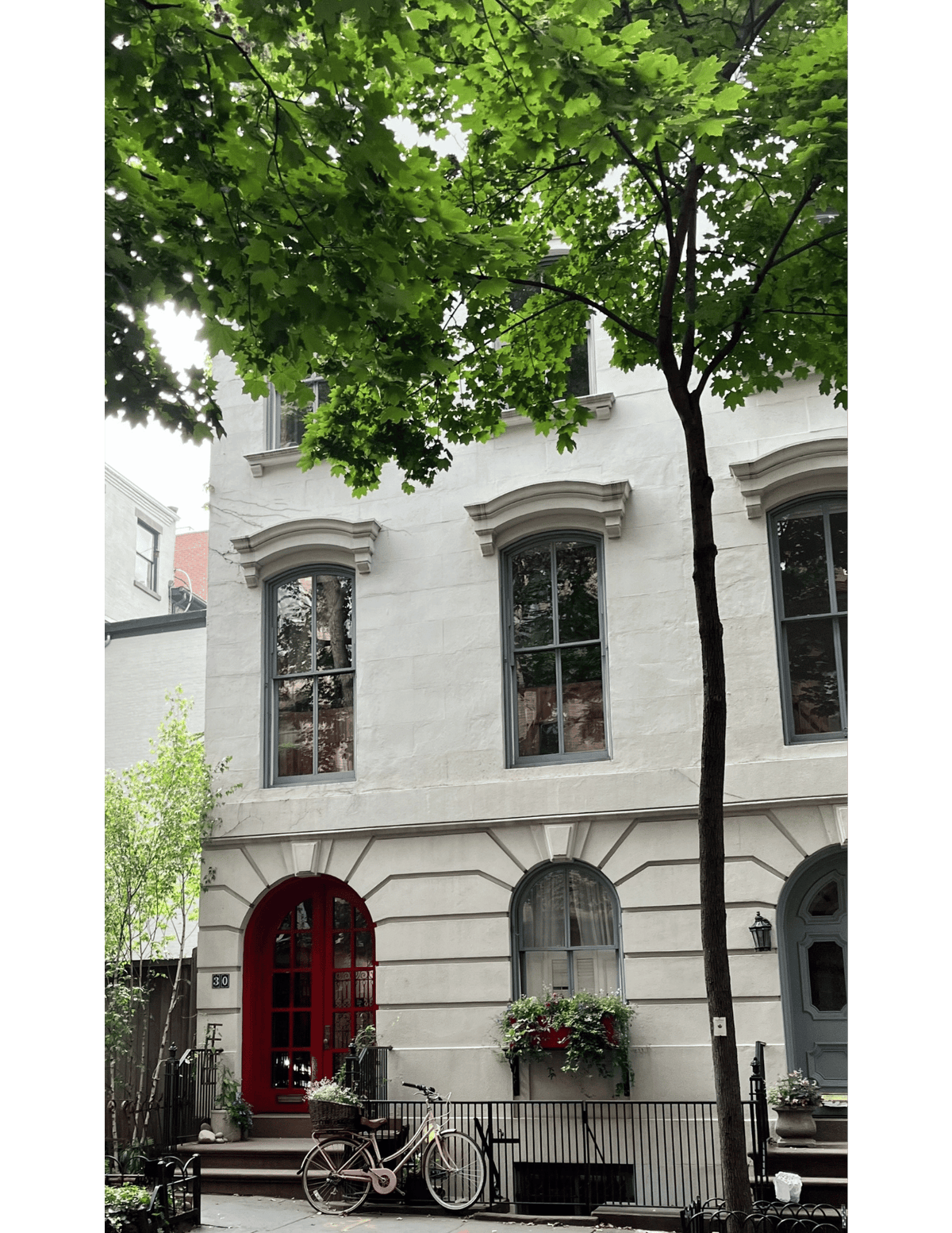 A white stone townhouse facade in Brooklyn, New York, with gray-framed windows, a red arched front door, a blue-gray side door, flower boxes, a bicycle at the entrance, and leafy green trees in the foreground.