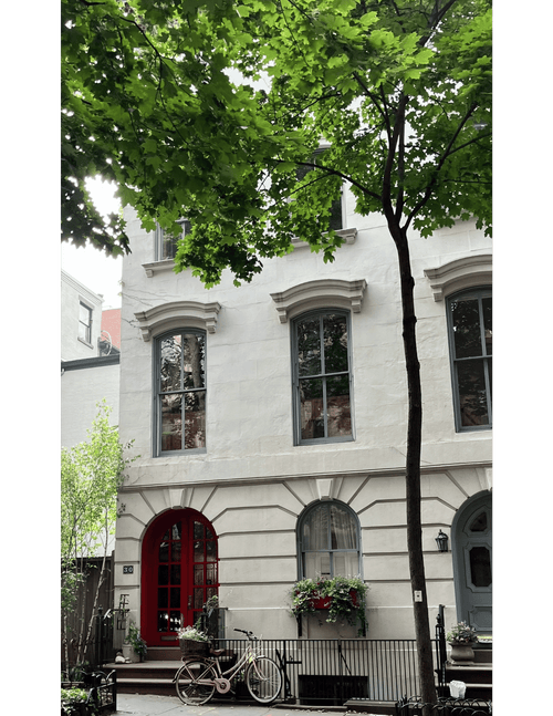 A white stone townhouse facade in Brooklyn, New York, with gray-framed windows, a red arched front door, a blue-gray side door, flower boxes, a bicycle at the entrance, and leafy green trees in the foreground.