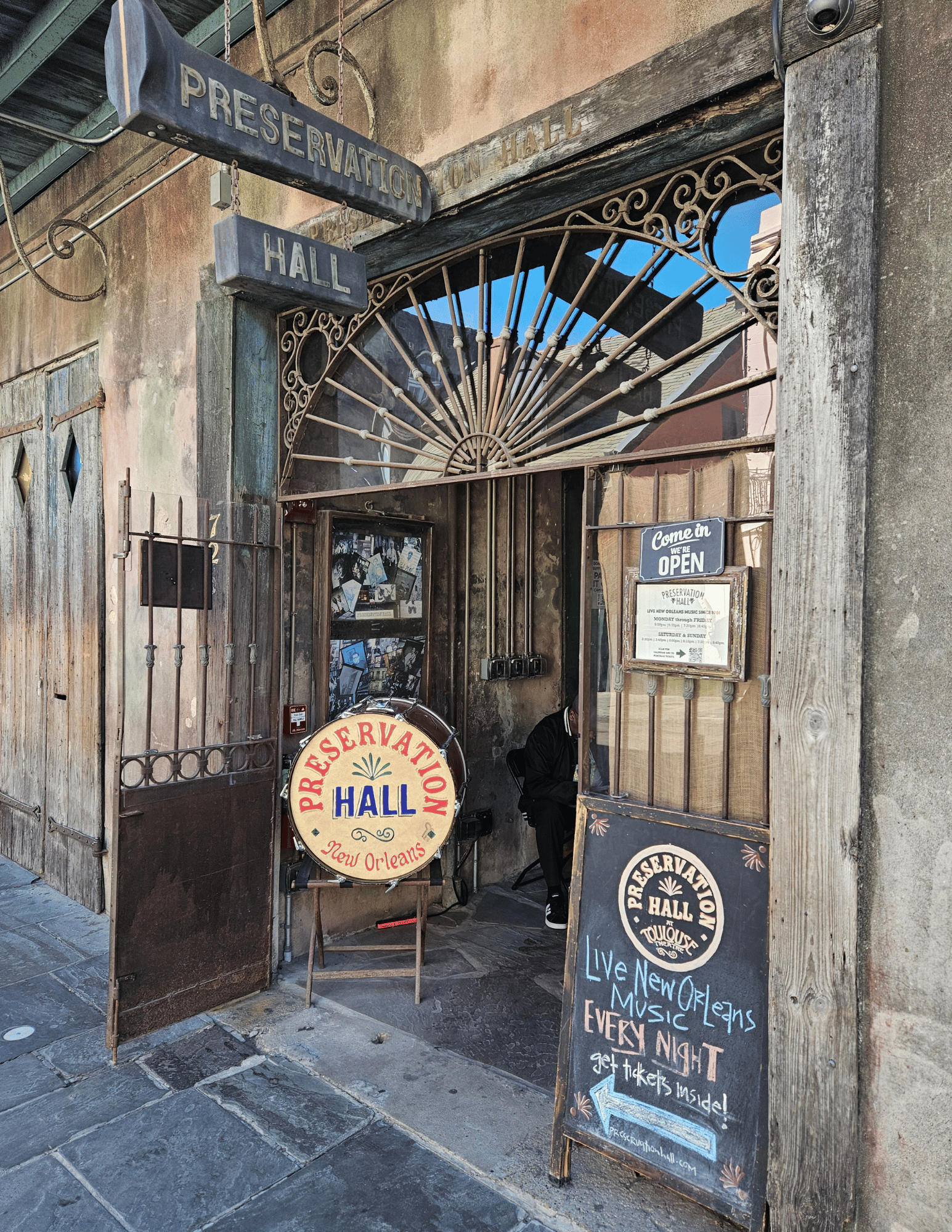 The entrance to Preservation Hall, a historic music venue in New Orleans, featuring ornate metal fan grates, rustic wooden doors, a round wooden sign, and a chalkboard A-frame sign on a stone sidewalk.