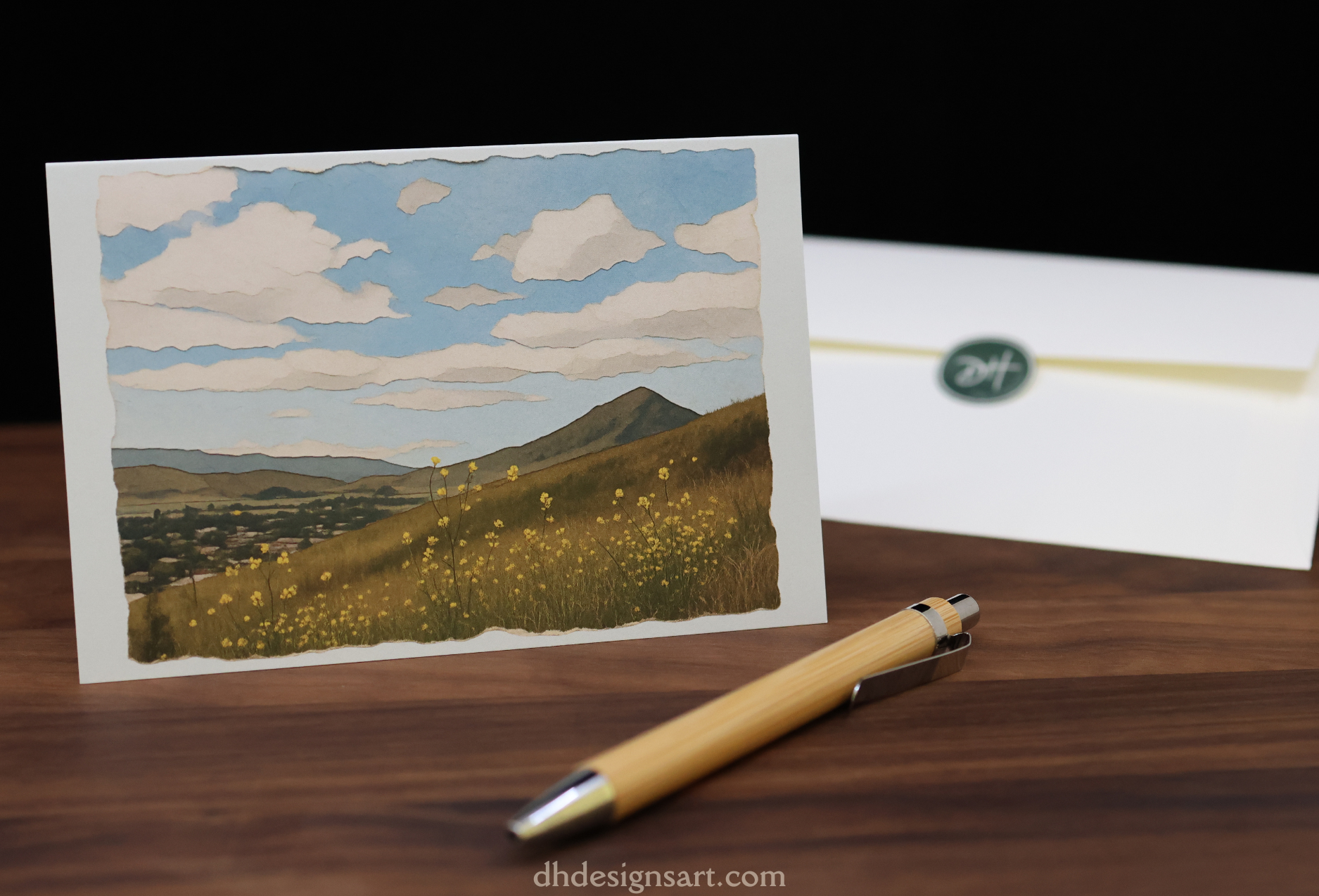 Blank notecard with a landscape design featuring mountains and fields, placed on a wooden desk with a pen - dhdesignsart.com.